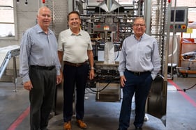Three men standing in front of an industrial bagging machine, from left to right is, David Muskat in a striped blue button up shirt, then Paul Muskat in an off-white polo, then Bryan Muskat in a grey/blue button up.