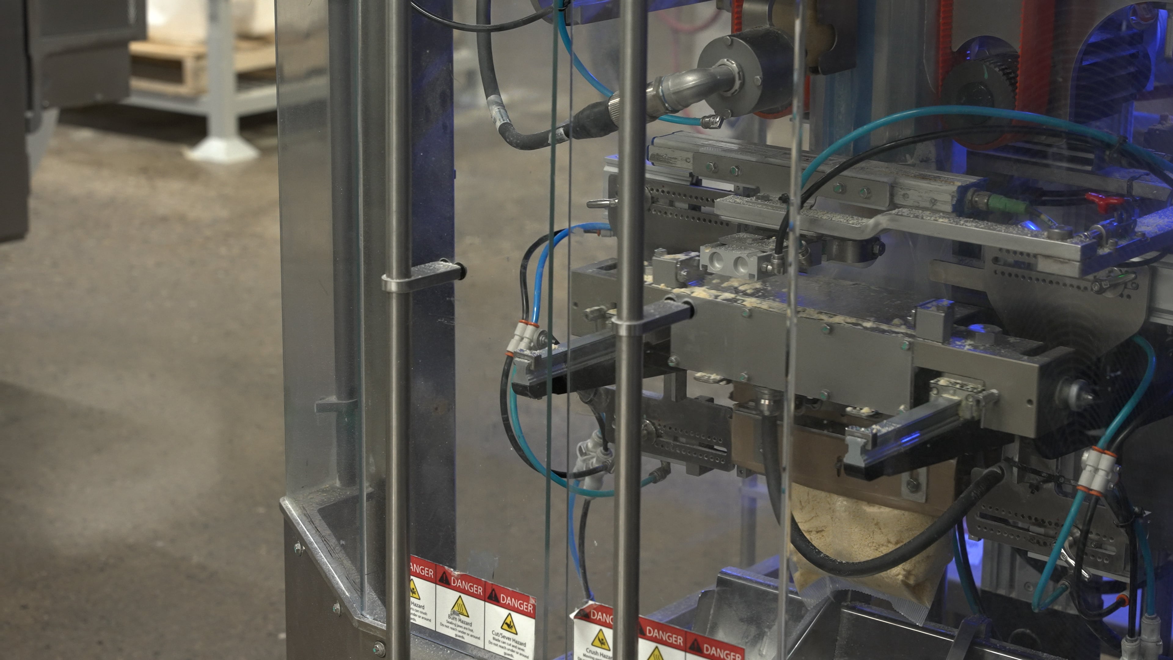 Close-up of a vertical form fill seal (VFFS) bagging machine packaging powder product into a flexible pouch. The image shows sealing components, pneumatic tubing, sensors, and stainless steel machine elements enclosed within a safety guard during automated packaging operation.